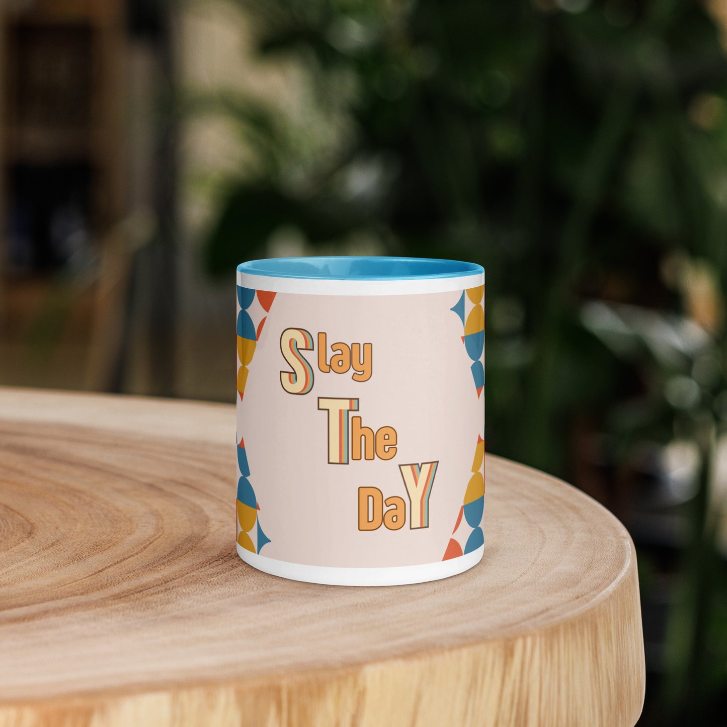 A yellow ceramic mug, named the "Slay the Day Mug," with "Slay The Day" inscribed on it and adorned with a geometric pattern, rests on a wooden table. It is both dishwasher and microwave safe for ultimate convenience.