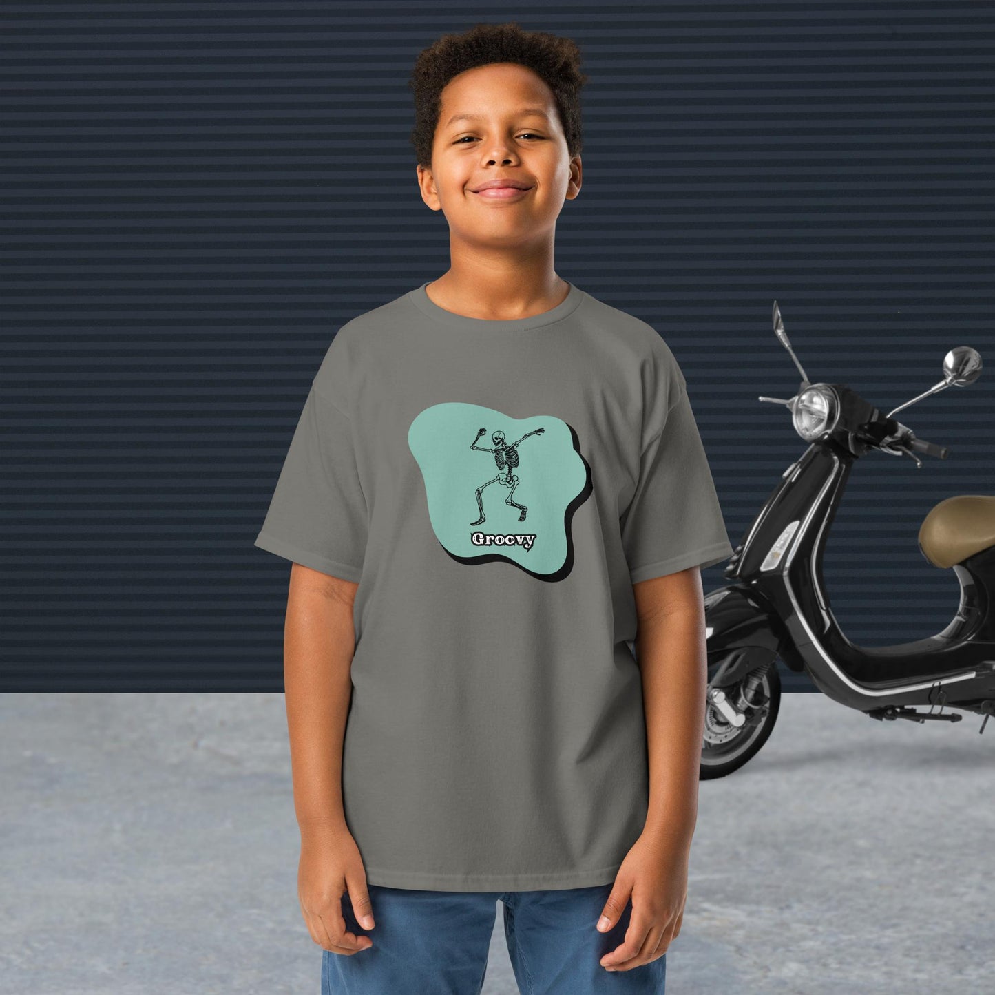 A boy wearing a royal Groovy Skeleton Design - Youth classic tee stands in front of a black scooter and a textured dark wall.