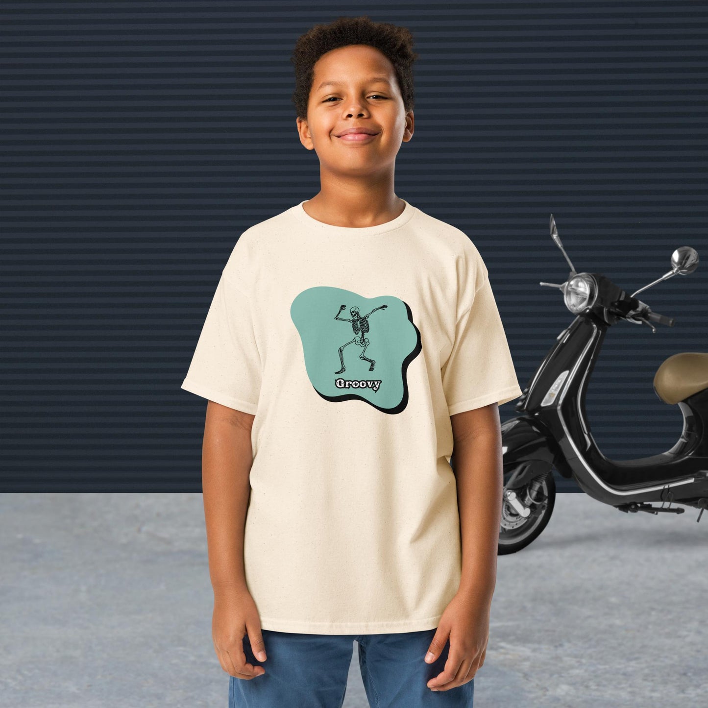 A boy wearing a royal Groovy Skeleton Design - Youth classic tee stands in front of a black scooter and a textured dark wall.