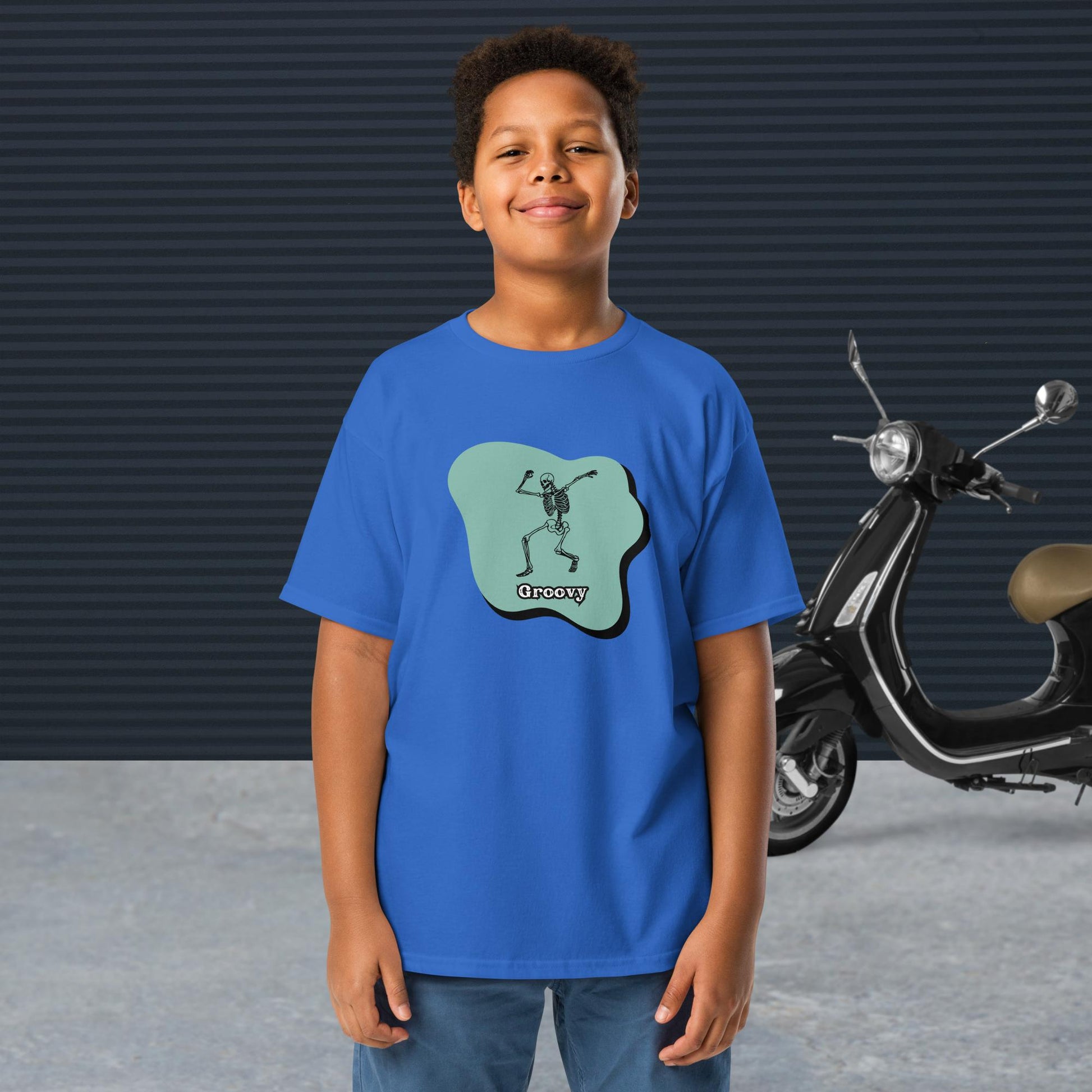 A boy wearing a royal Groovy Skeleton Design - Youth classic tee stands in front of a black scooter and a textured dark wall.