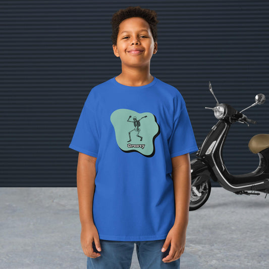 A boy wearing a royal Groovy Skeleton Design - Youth classic tee stands in front of a black scooter and a textured dark wall.